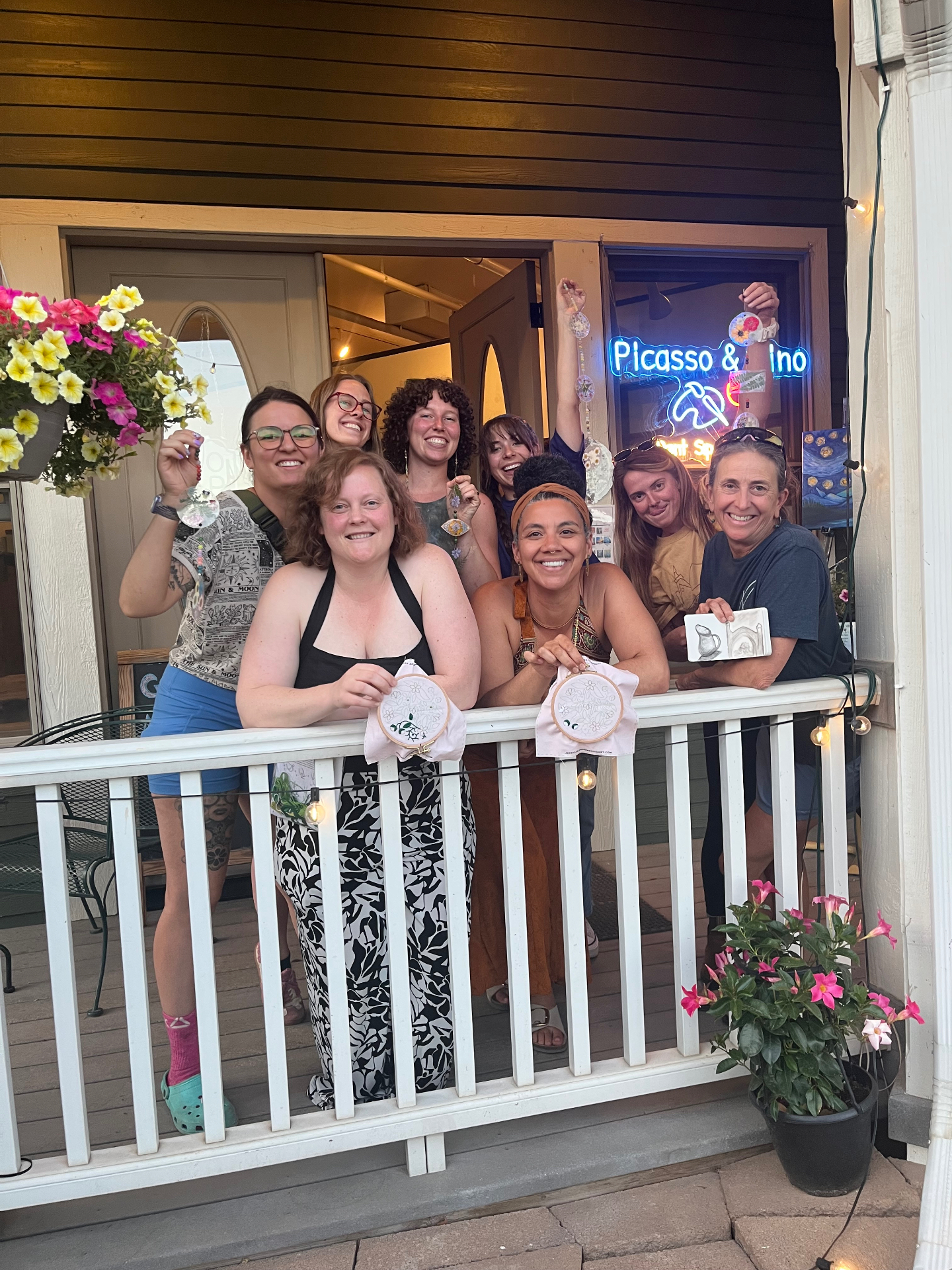 A joyful group on the Rabbitbrush Studio porch showing off their handmade embroidery hoops and pressed-flower suncatchers by the Picasso & Vino neon sign